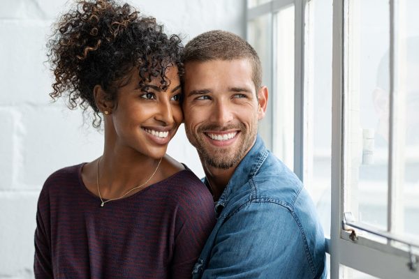 Loving multiethnic couple embracing and sitting near window. Happy girlfriend and smiling boyfriend looking away and thinking about their future together. Young man and african woman contemplating.
