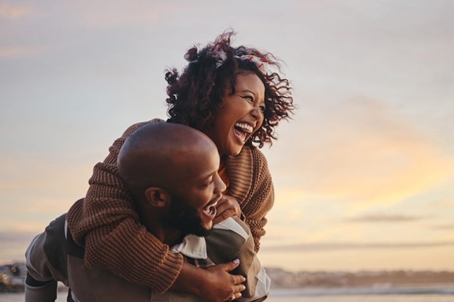 Black couple, travel and beach fun while laughing on sunset nature adventure and summer vacation or honeymoon with a piggy back ride. Comic, energy and black man and woman love on ocean holiday.